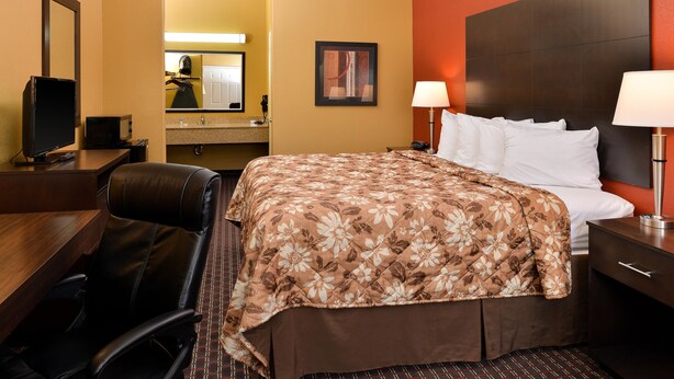 A hotel room with a bed, a desk, a chair, a television, a mirror, and a bathroom visible through an archway. - Americas Best Value Inn Tupelo (Tupelo)
