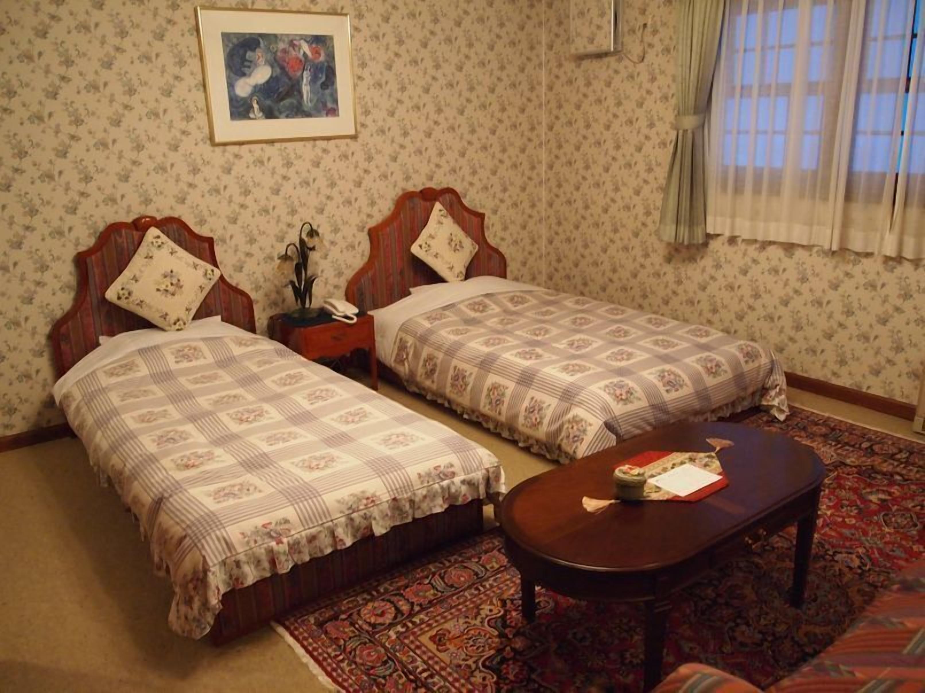 A room with two beds, a nightstand, a table, a patterned rug, and a framed picture on the wall.