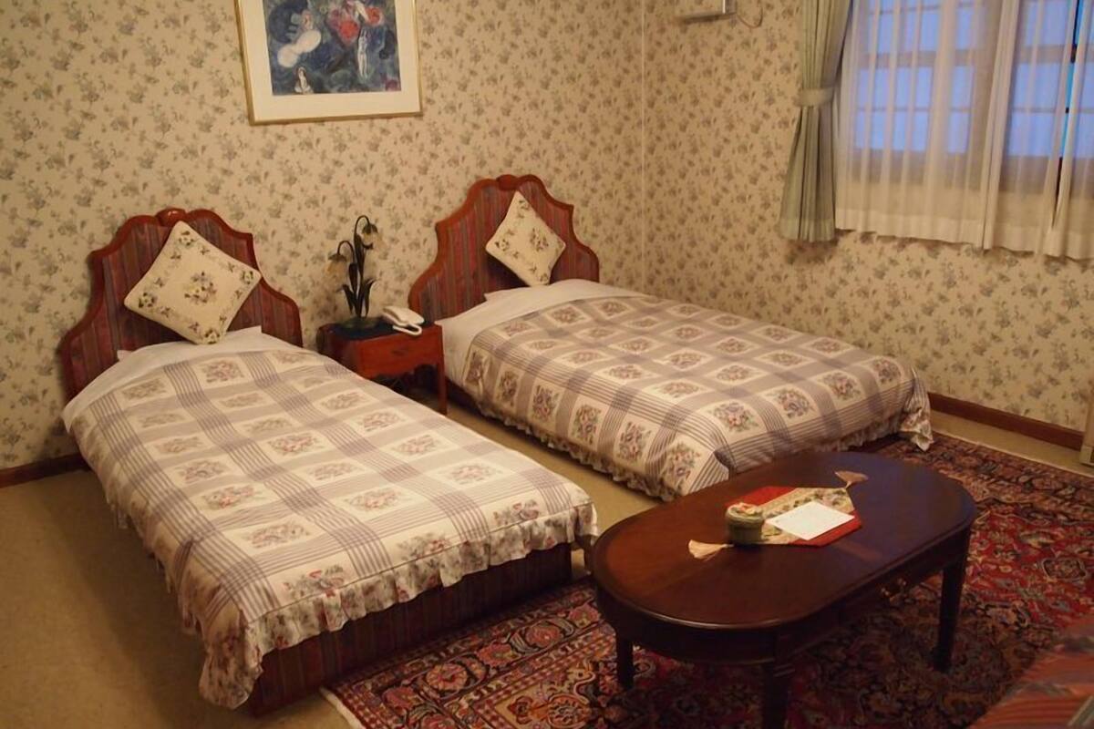 Une chambre avec deux lits, une table de chevet, une table, un tapis à motifs et un tableau encadré au mur.