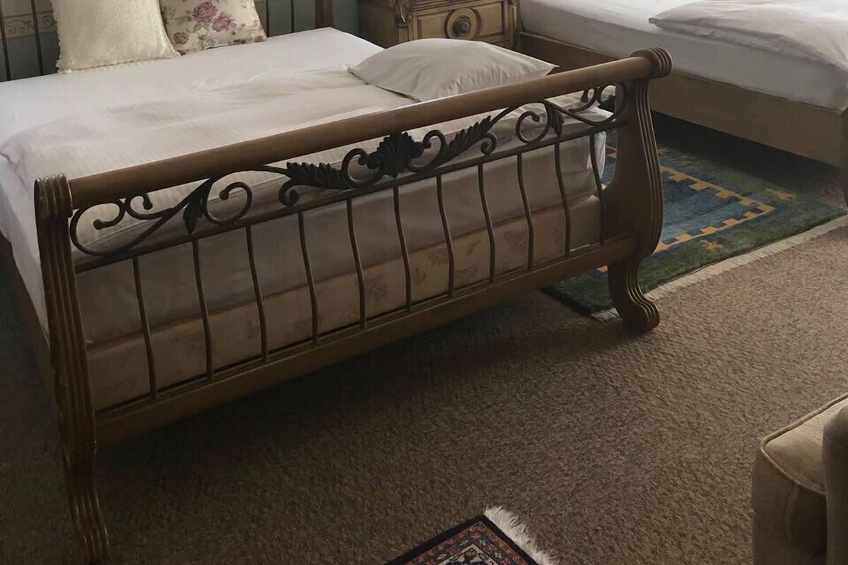 Deux lits jumeaux dotés de têtes de lit en métal décorées, une petite table de chevet avec une lampe et une fenêtre agrémentée de rideaux à motifs floraux.