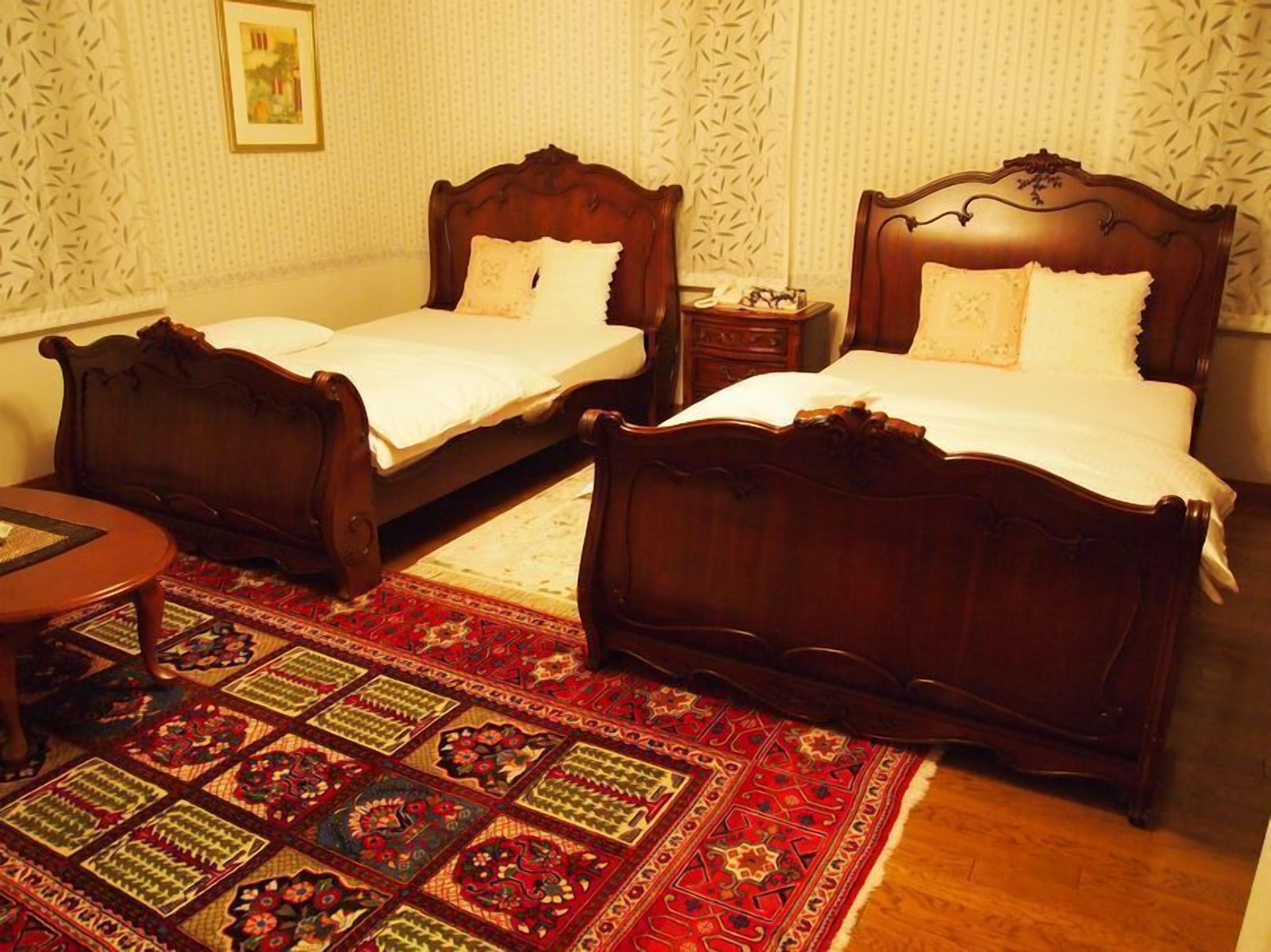 A hotel room with two wooden beds, a small table, and a patterned rug.