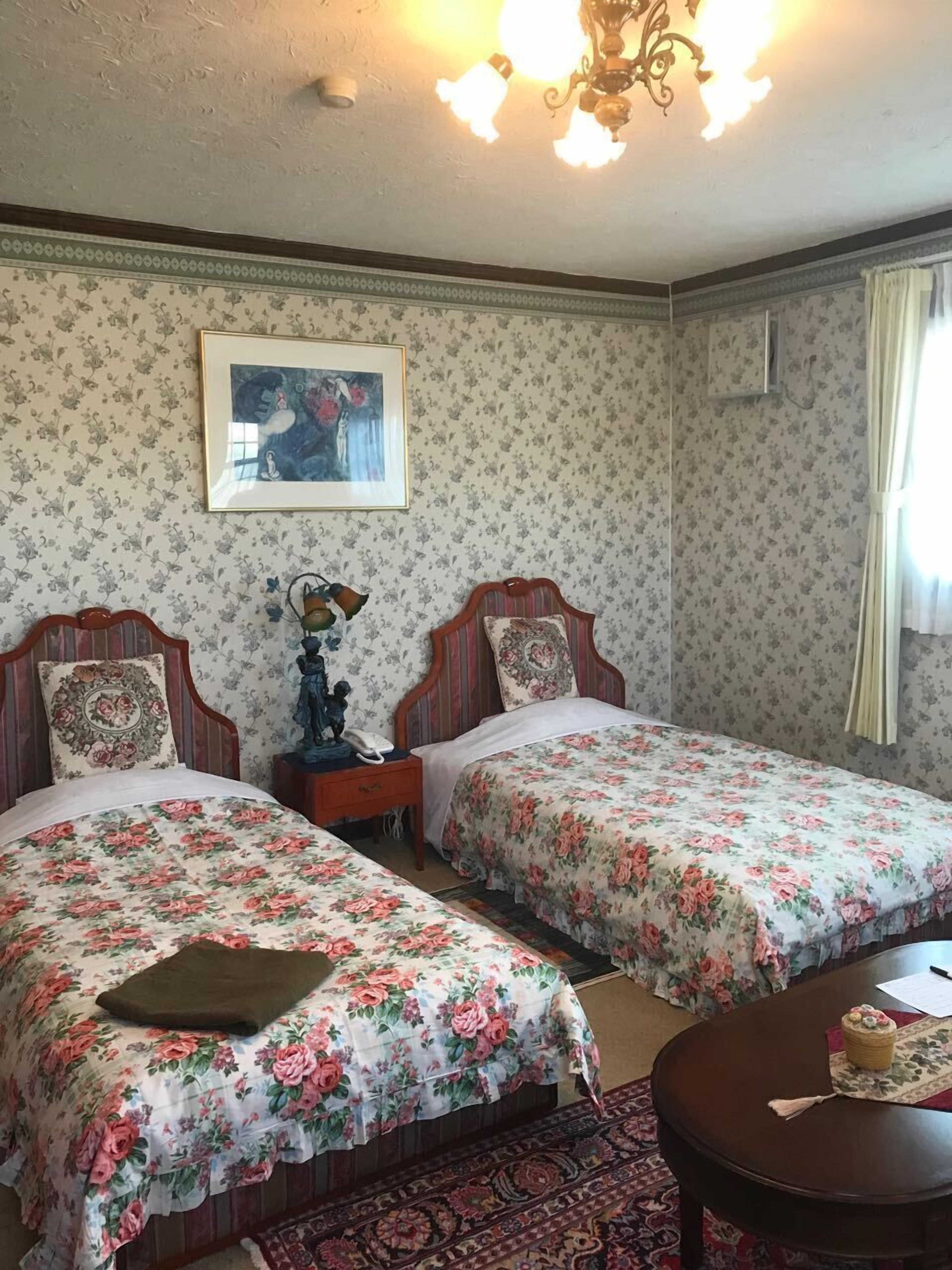 A room with two beds, a nightstand, a framed picture, a chandelier, and a small table with a basket.