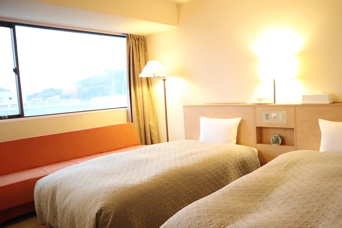 A hotel room with two beds, a nightstand, a lamp, a clock, and a window with curtains.