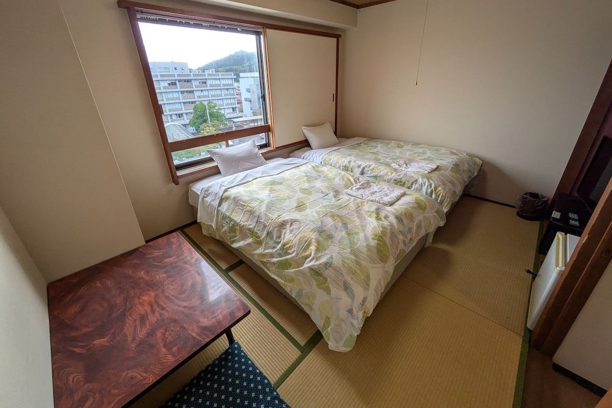 A small room with a bed, a wooden desk, and a window with a view of buildings.