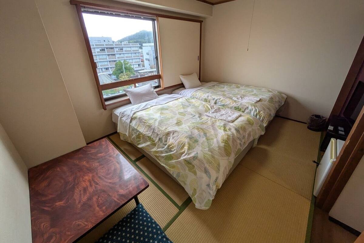 A small room with a bed, a wooden desk, and a window with a view of buildings.