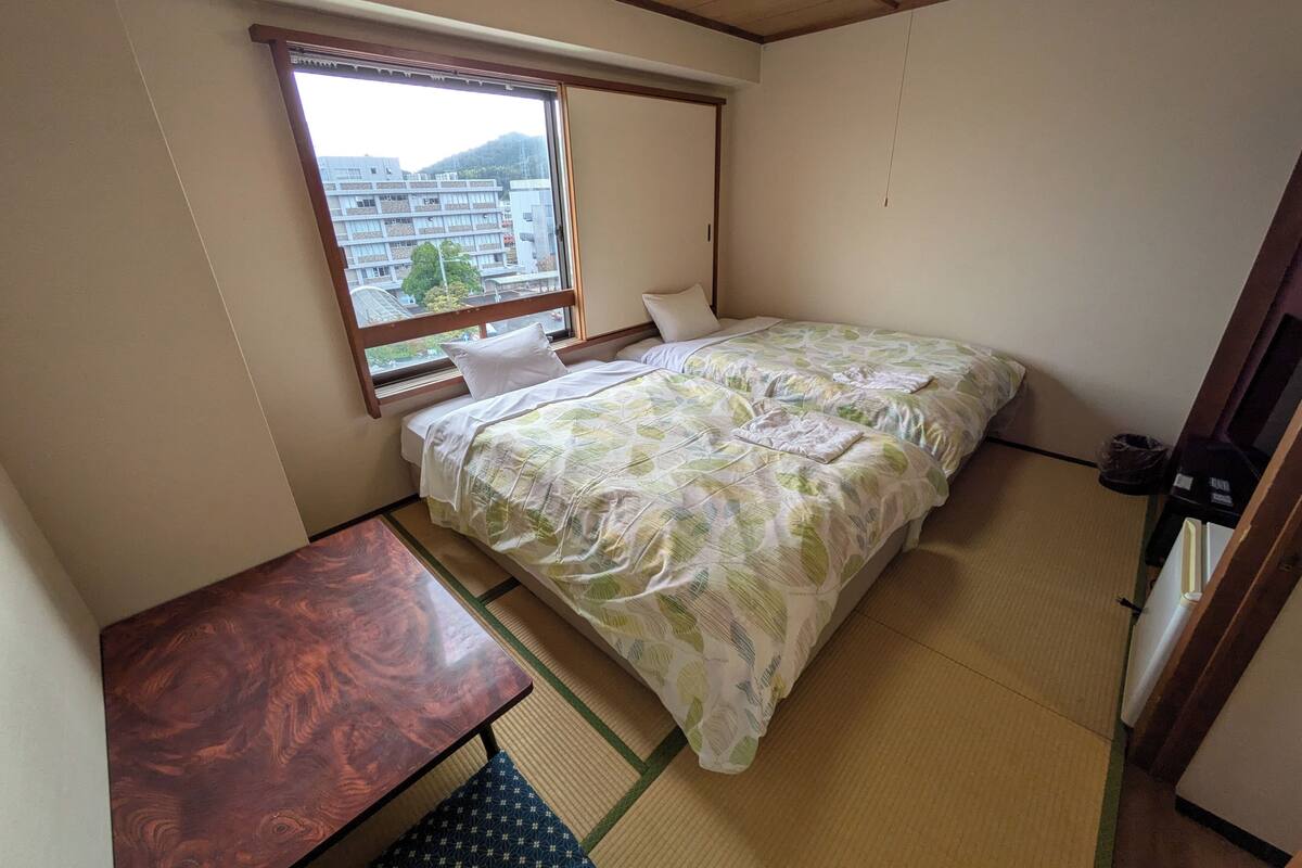 A room with a bed, a small table, a window with a view of buildings and trees, and a wooden ceiling.