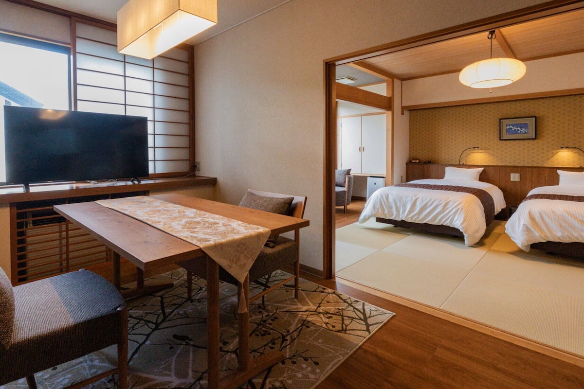 A hotel room with a TV, a dining table, two beds, and a view of the outside.