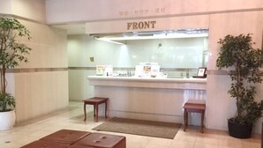 A reception area with a counter, two wooden stools, a brown cushioned bench, and potted plants. - Hotel Hitachi Plaza (Hitachi)