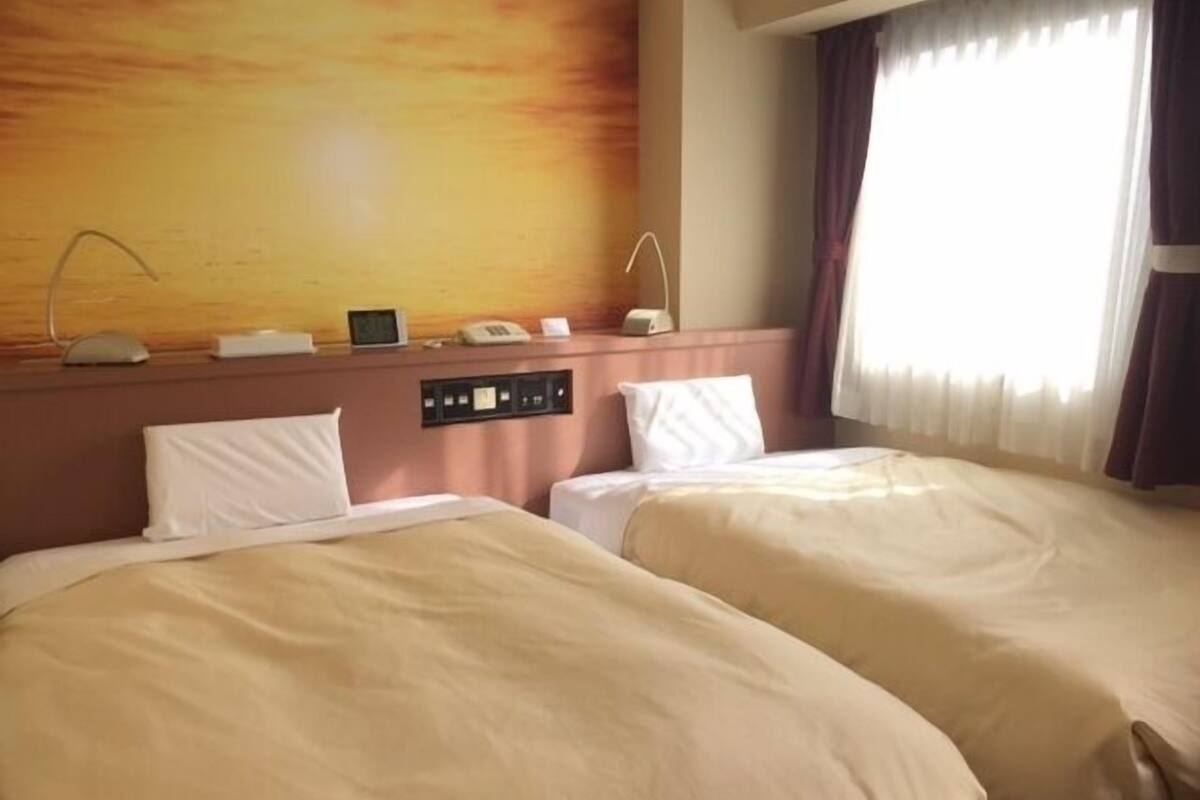 Une chambre d’hôtel avec deux lits, un panneau de contrôle, une horloge et une fenêtre avec des rideaux.