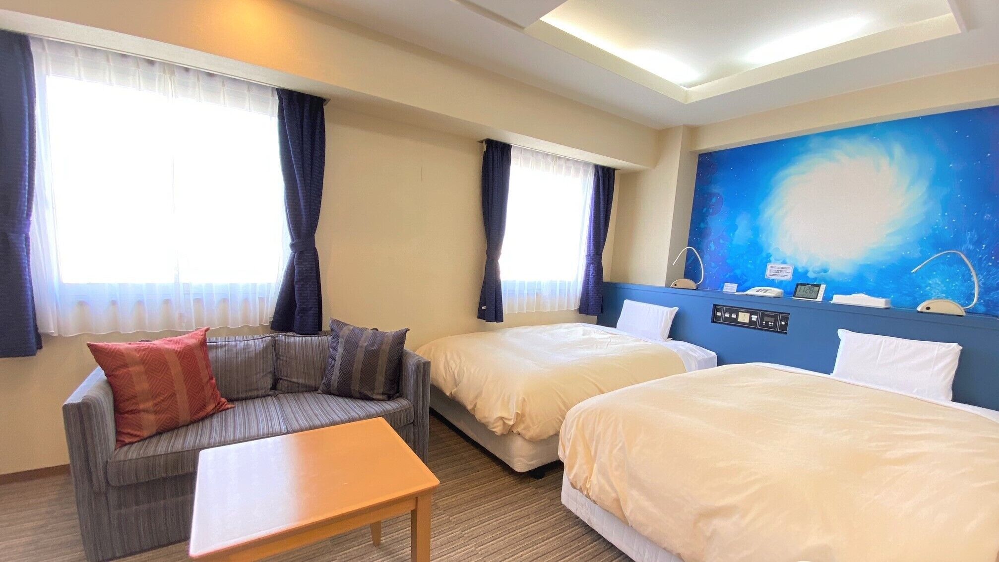A hotel room with two beds, a sofa, a table, and a large window with curtains.
