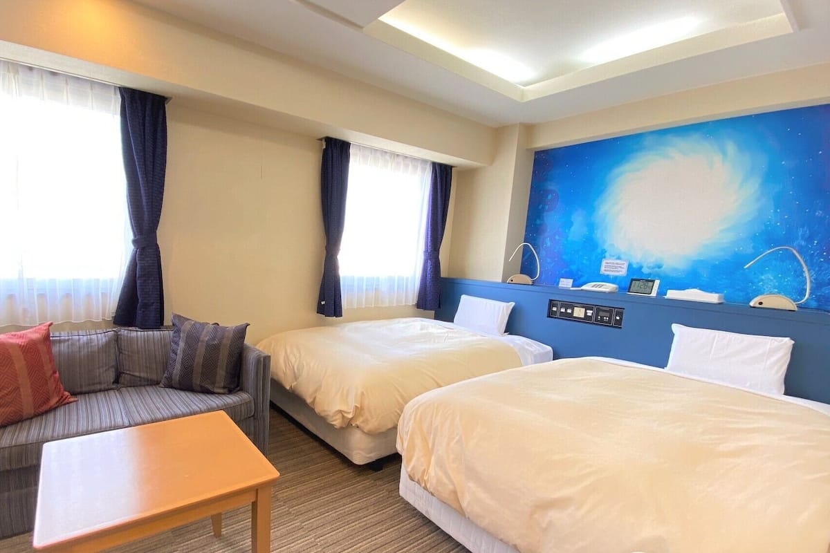 Une chambre d’hôtel avec deux lits, un canapé, une table et une grande fenêtre avec des rideaux.