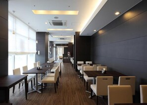 A modern hotel lobby with long tables and chairs, a wooden floor, and a dark wall on one side. - Hotel Sunroute Fukuchiyama (Fukuchiyama)