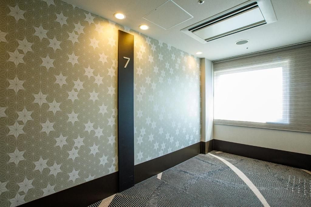 A hotel room with a patterned wall, a number sign, a window with blinds, and a carpeted floor.