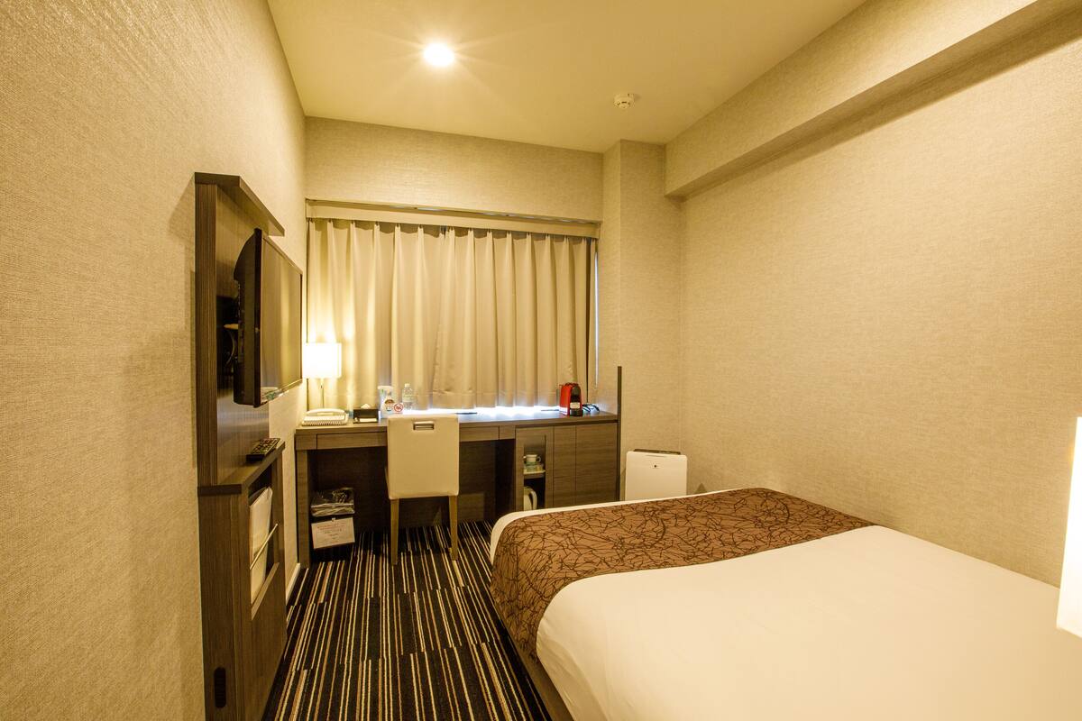A hotel room with a bed, a desk with a chair, a television, and a window with curtains.