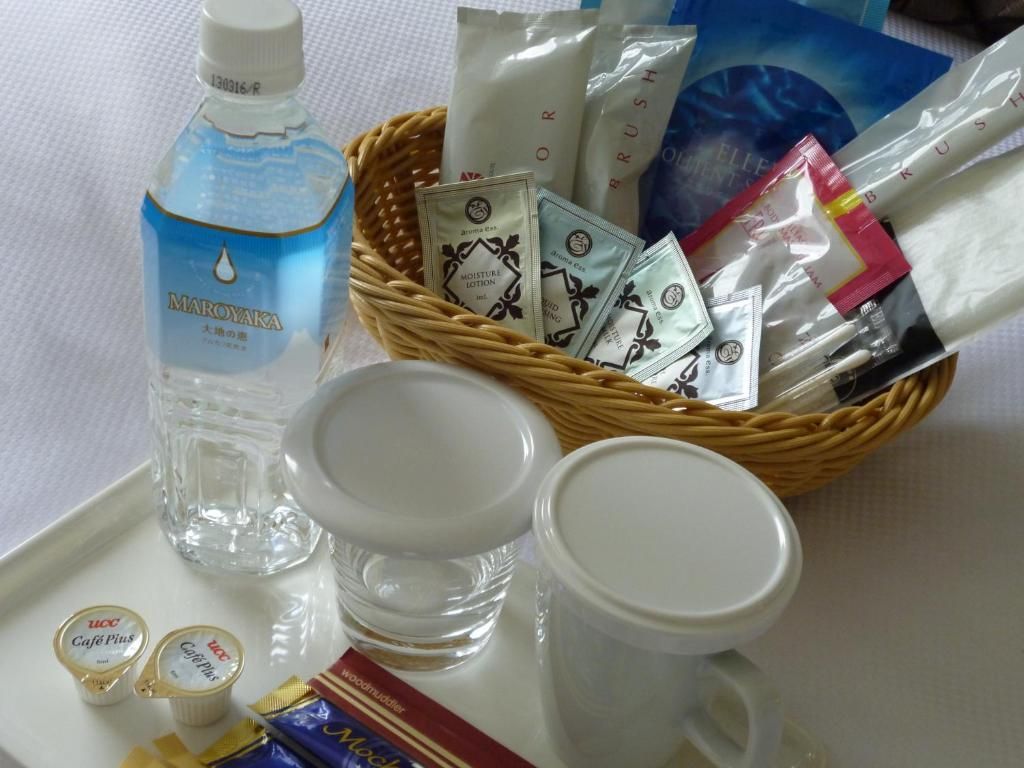 Una bandeja con una botella de agua Maroyaka, un cesto de artículos de aseo y dos tazas blancas vacías.