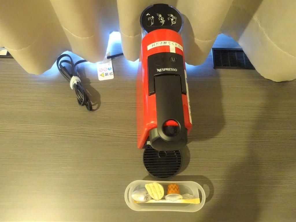 Une machine à café Nespresso rouge, avec un compartiment pour les capsules de café et un câble d’alimentation, posée sur une surface en bois.