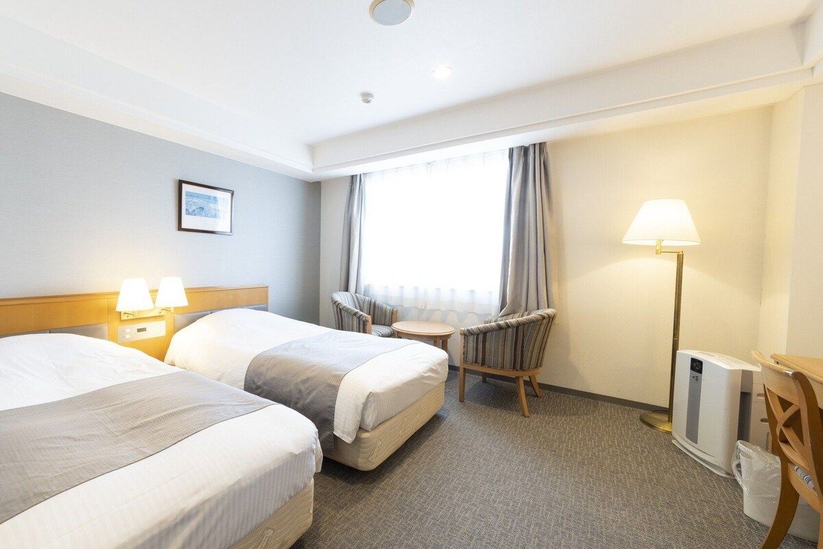 A hotel room with two beds, a chair, a small table, a lamp, and a window with curtains.