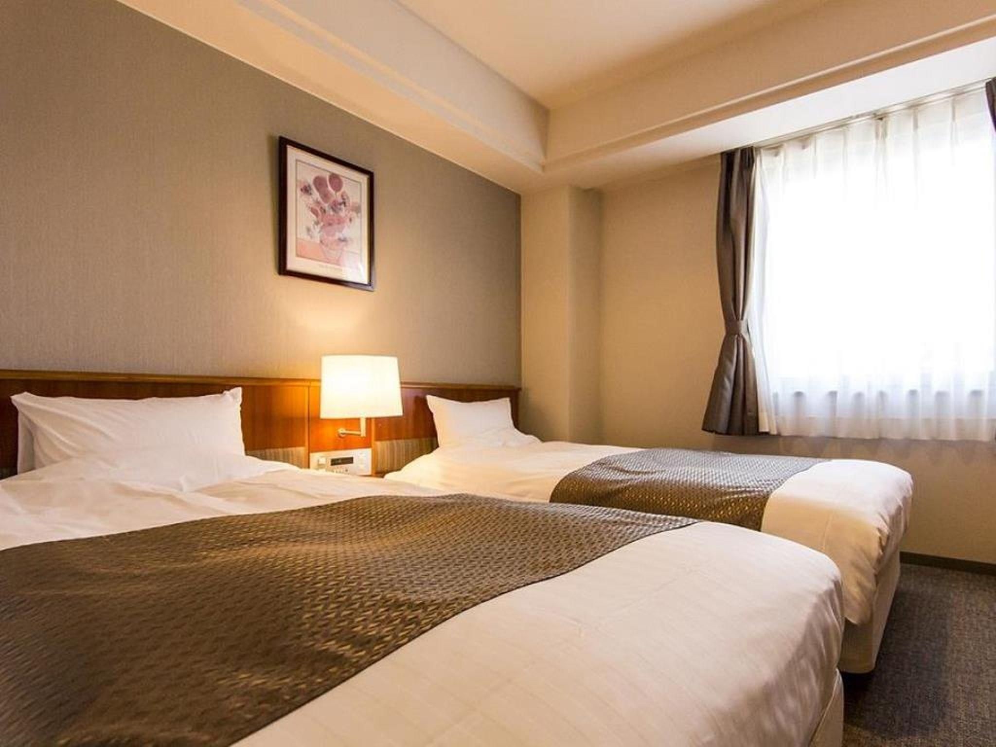 Habitación de hotel con dos camas, una mesita de noche con lámpara, un cuadro enmarcado en la pared y una ventana con cortinas.