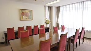 A conference room with a long table and red chairs, a painting on the wall, and a window with curtains.