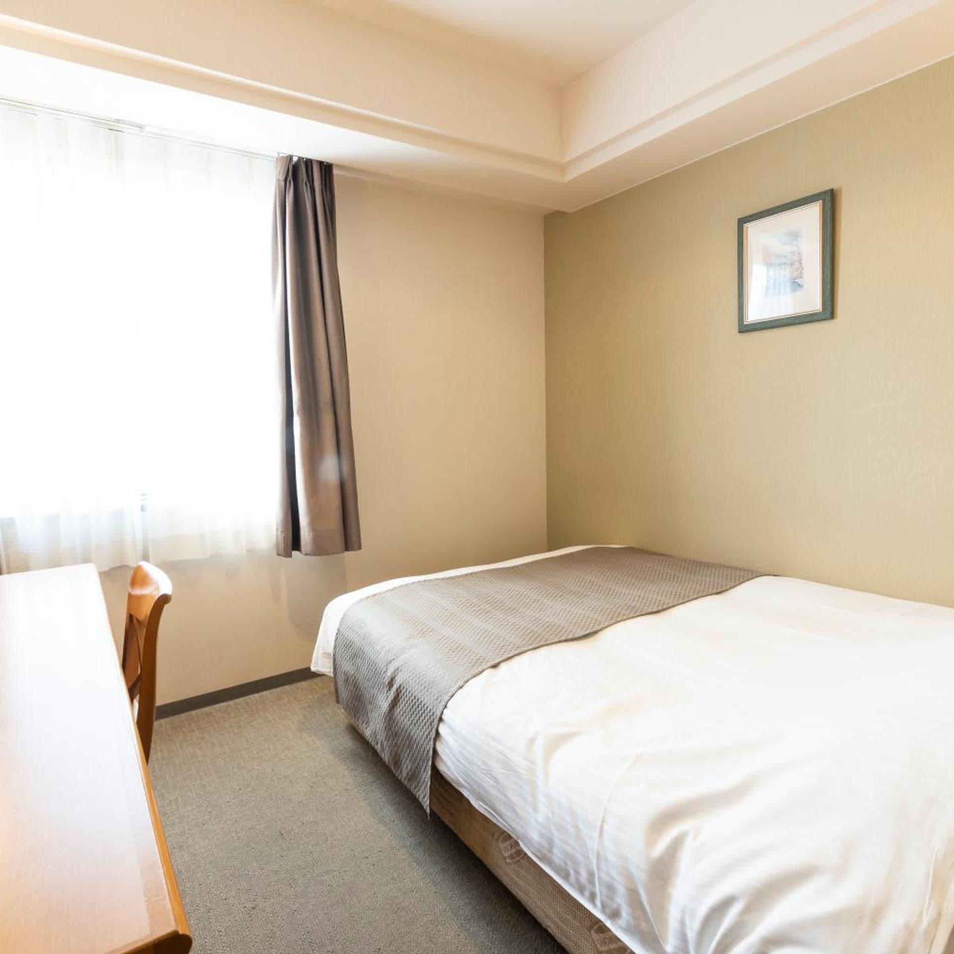 A hotel room with a bed, a desk, a chair, a window with curtains, and a framed picture on the wall.