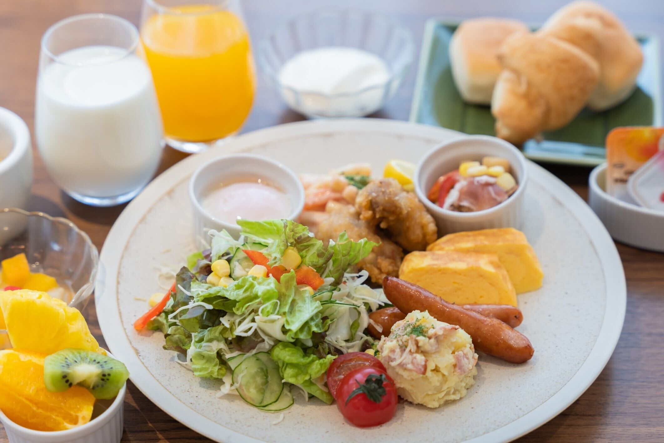 A plate of food including a salad, grilled chicken, sausages, and cheese, with drinks and bread in the background.