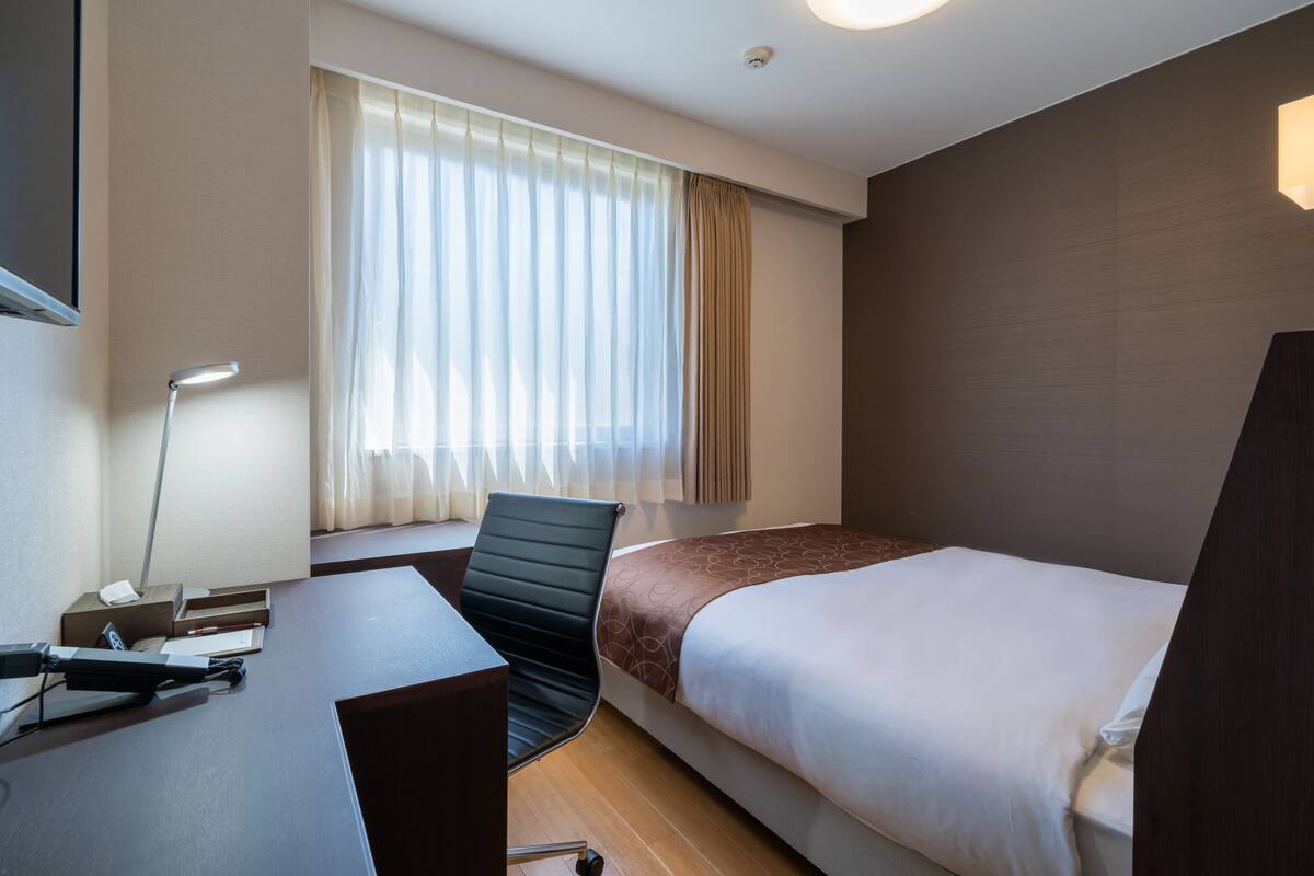 Hotelkamer met een bed, bureau, stoel, raam met gordijnen en een wandlamp.