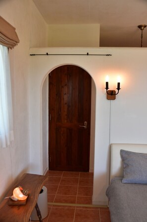 A room with a wooden door, a bed with a grey blanket, a wall-mounted light fixture, and a small table with a candle.