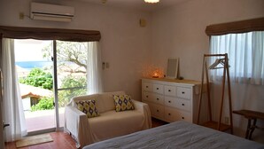 A bedroom with a bed, a sofa, a dresser, and a window with a view of greenery and a building. - Terrace House Yamabare (Ishigaki)