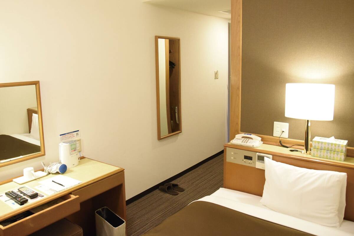 Una habitación de hotel con una cama, un escritorio con teléfono y lámpara, un espejo y un cesto de basura.