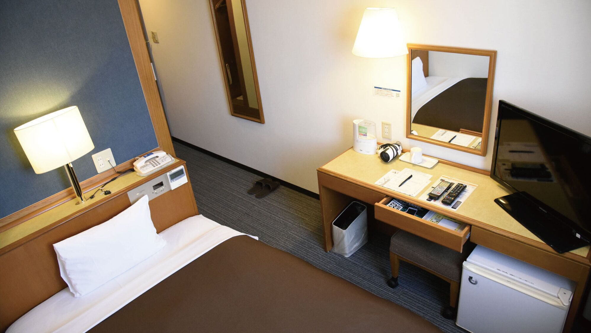 A hotel room with a bed, a desk with a lamp, a mirror, a TV, and a trash bin.