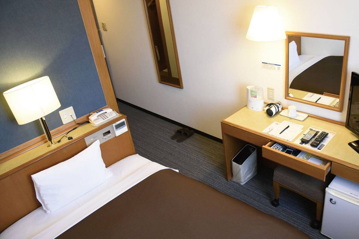 Una habitación de hotel con una cama, un escritorio con lámpara, un espejo, un televisor y un cesto de basura.