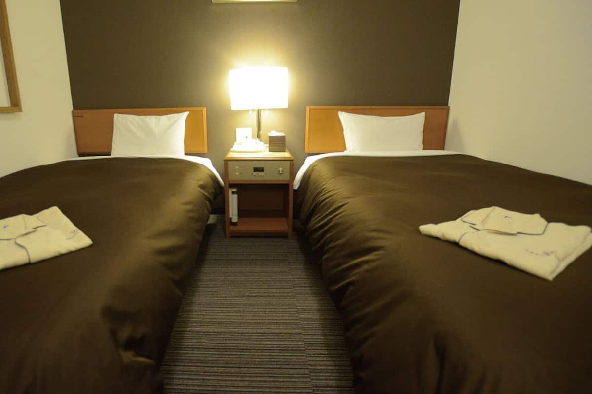 Habitación de hotel con dos camas, una mesita de noche, una lámpara y piso alfombrado.