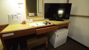 Une chambre d’hôtel équipée d’un bureau en bois, d’une télévision, d’un mini-réfrigérateur, d’un miroir et d’une petite table de chevet avec une lampe.