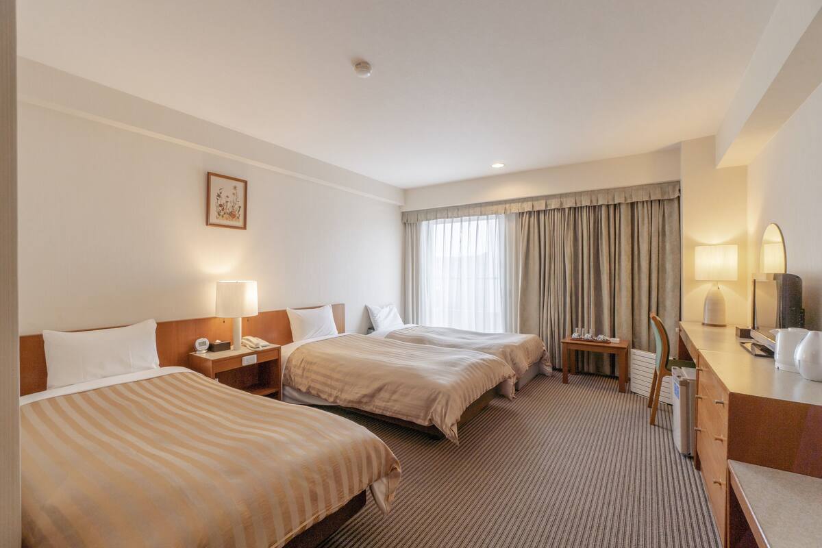 Ein Hotelzimmer mit zwei Betten, einem Schreibtisch mit Lampe, einem Stuhl, einem kleinen Tisch und einem Fenster mit Vorhängen.