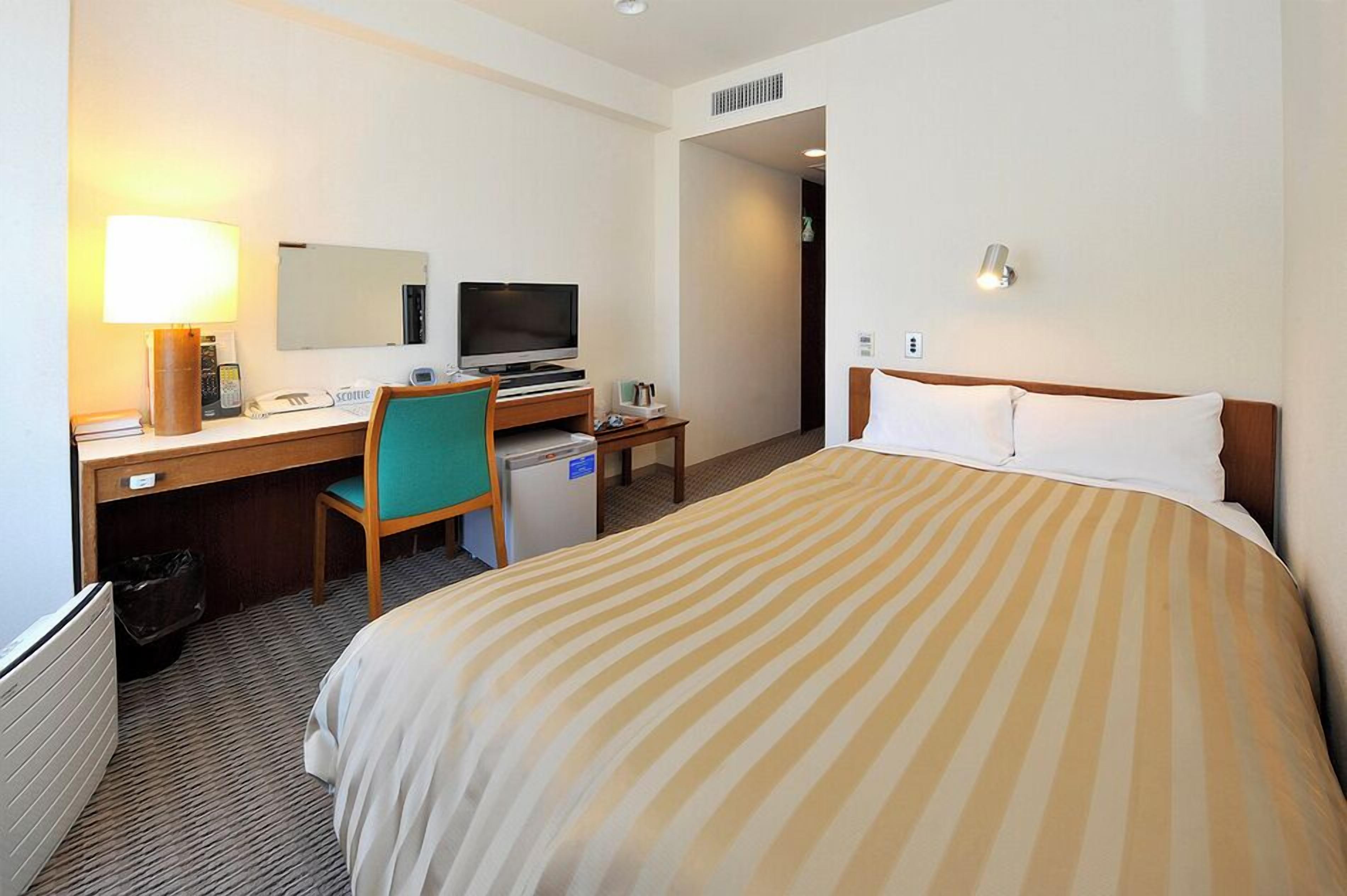 A hotel room with a bed, desk, chair, television, and a small kitchenette.