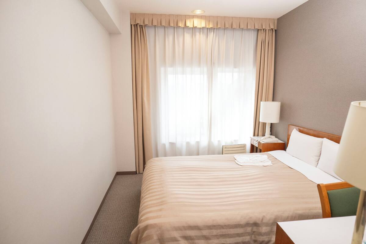 Ein Hotelzimmer mit einem großen Bett, einem Nachttisch, einer Lampe und einem Fenster mit Vorhängen.