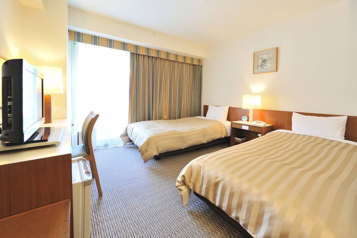 Ein Hotelzimmer mit zwei Betten, einem Schreibtisch mit Computer, einem Sessel, einer Schreibtischlampe, einem Nachttisch mit Lampe und einem Fenster mit Vorhängen.