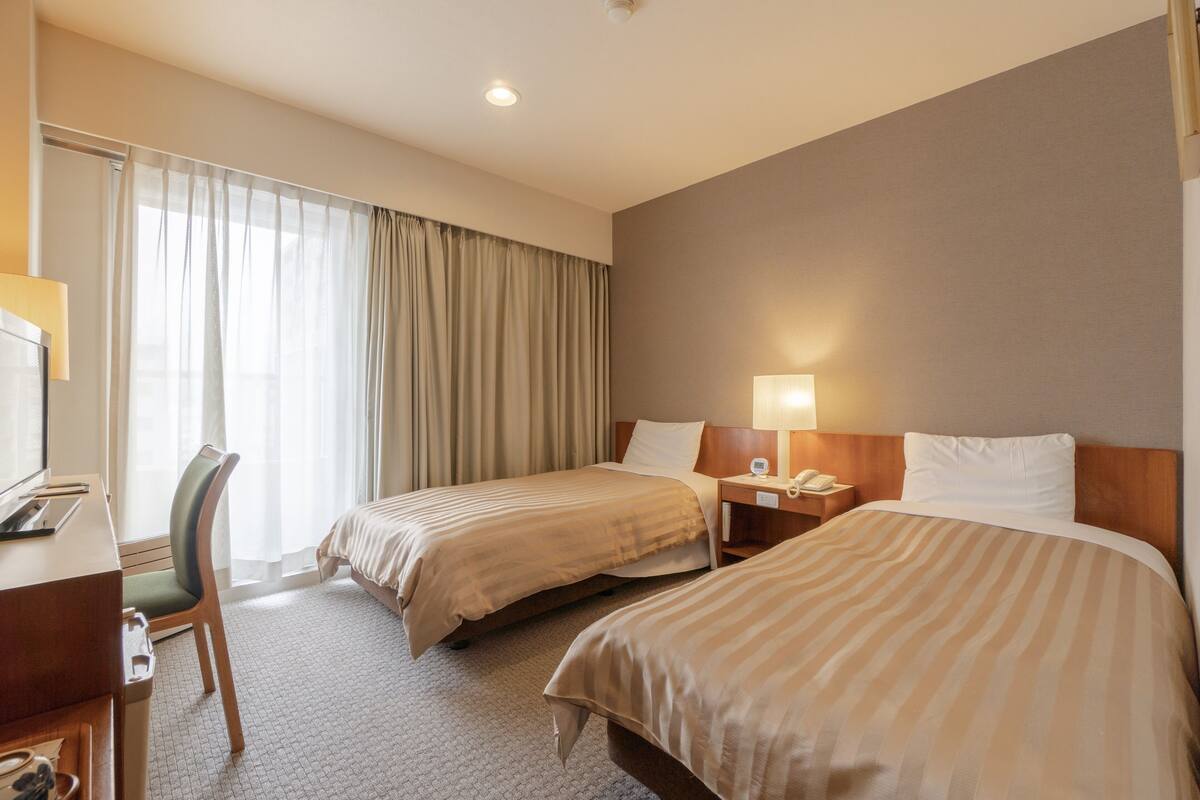 Ein Hotelzimmer mit zwei Betten, einem Schreibtisch, einem Stuhl und einem Fenster mit Vorhängen.
