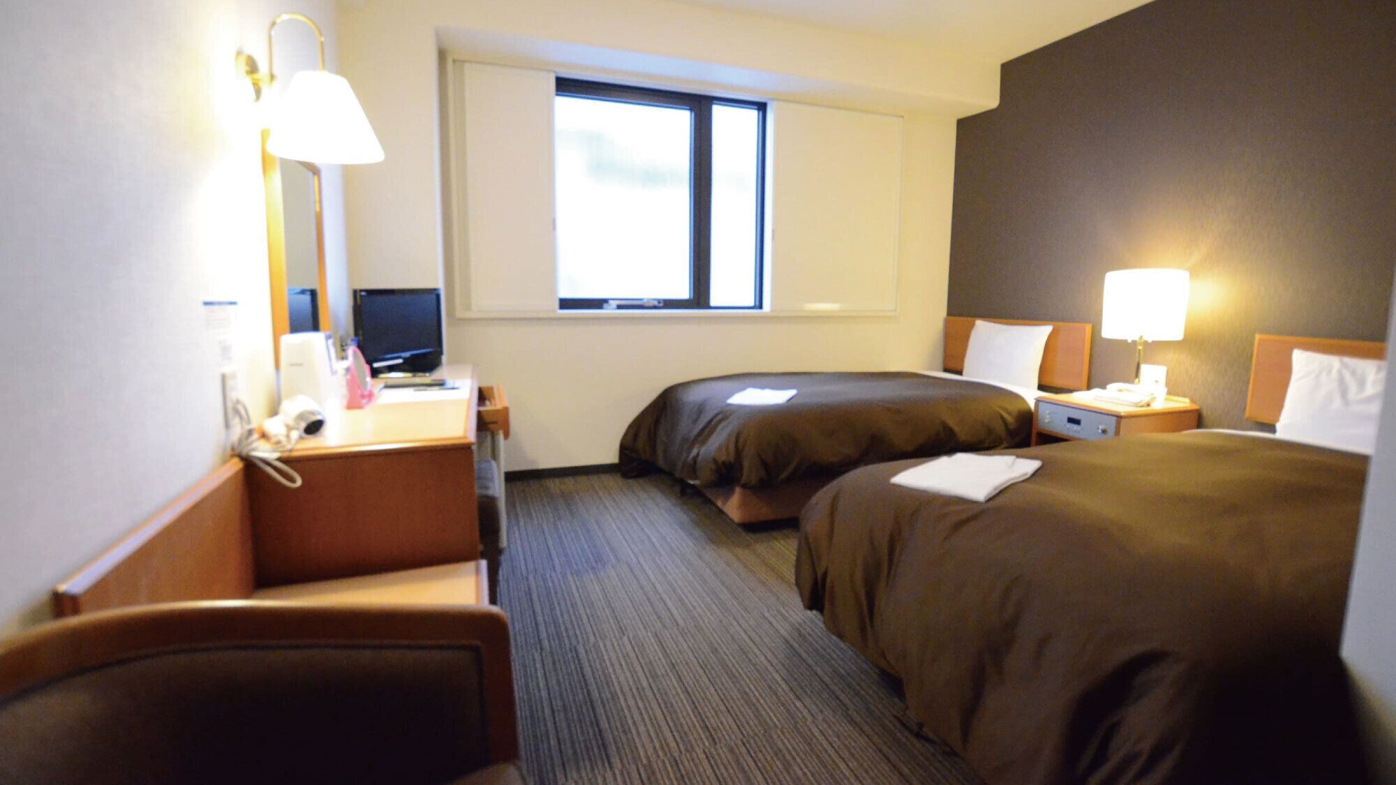 Hotellihuone, jossa on työpöytä, kaksi sänkyä, ikkuna ja yöpöytä, jossa on lamppu.