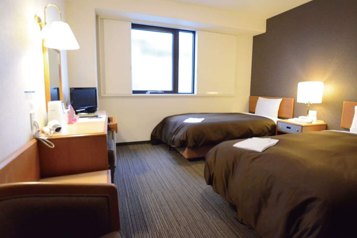 Une chambre d’hôtel équipée d’un bureau, de deux lits, d’une fenêtre et d’une table de chevet avec une lampe.