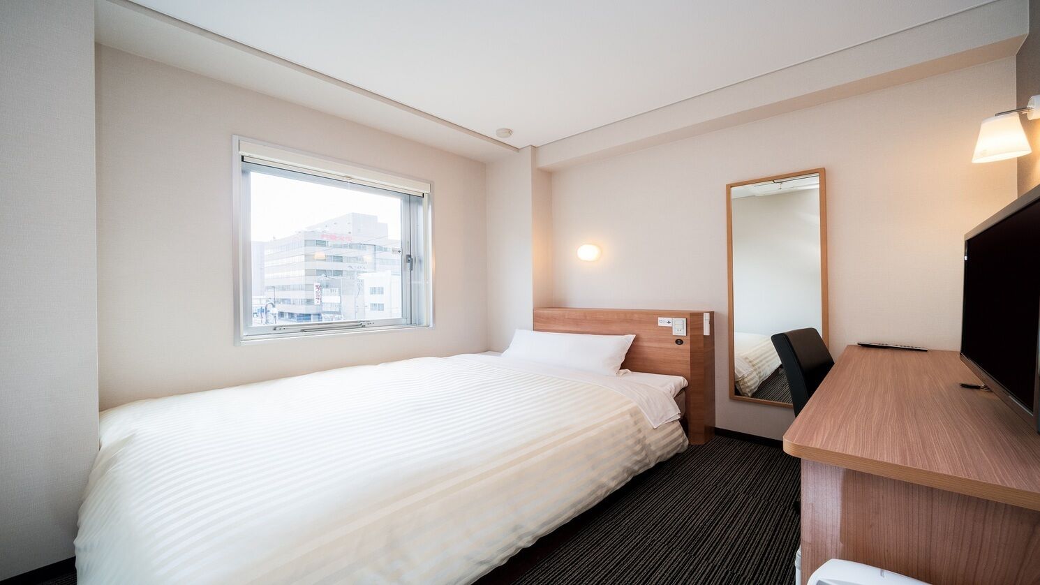 Une chambre d’hôtel avec un lit simple, un bureau, une chaise, un grand miroir et une fenêtre.