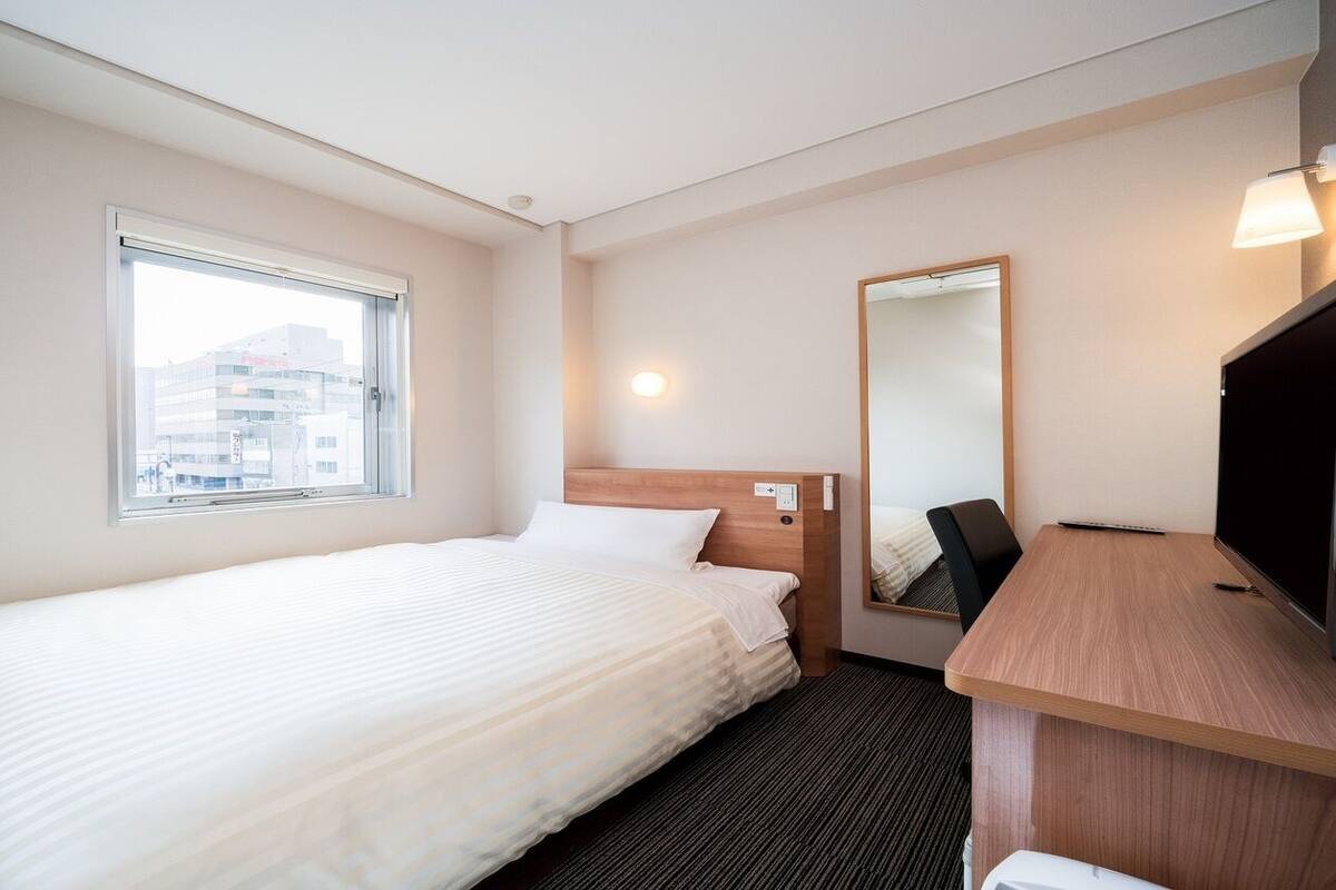 Une chambre d’hôtel avec un lit simple, un bureau, une chaise, un grand miroir et une fenêtre.