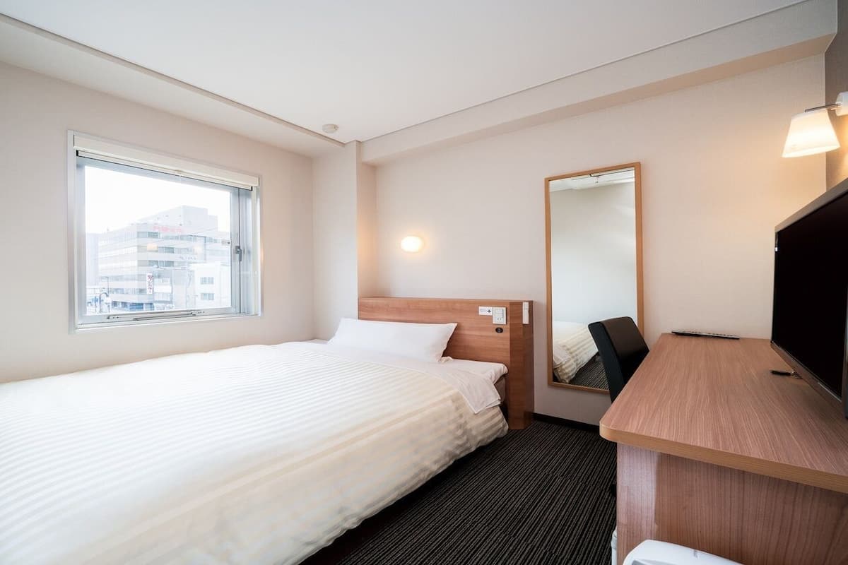 Une chambre d’hôtel avec un lit simple, un bureau, une chaise, un grand miroir et une fenêtre.