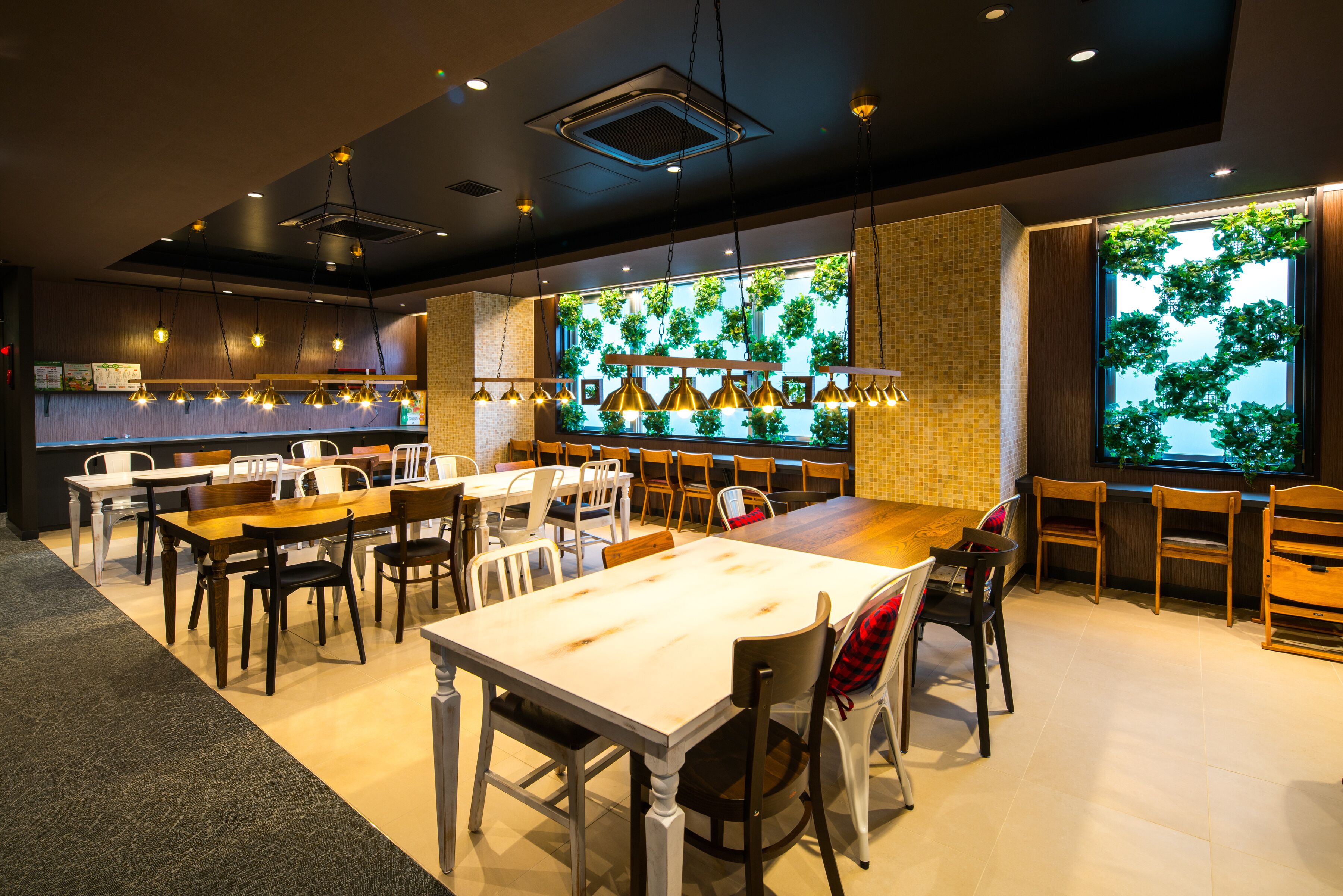 Un restaurante con una larga mesa de madera, varias sillas, lámparas colgantes y un amplio ventanal con vegetación.