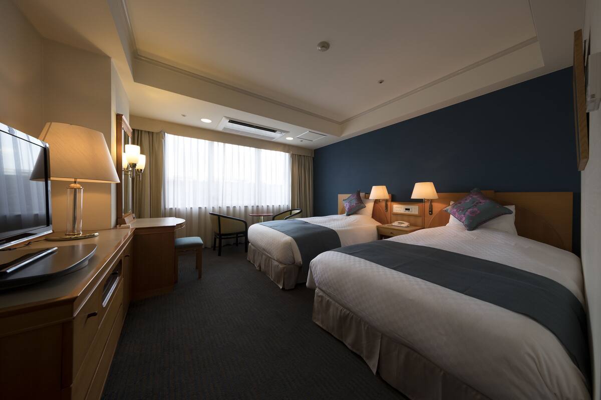 A hotel room with two beds, a desk, a chair, a TV, and a window with curtains.