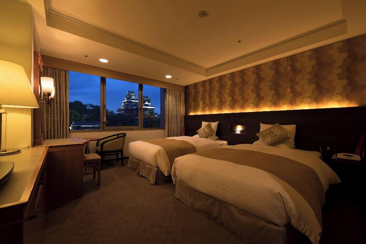 A hotel room with two beds, a desk, a chair, and a view of a castle through large windows.