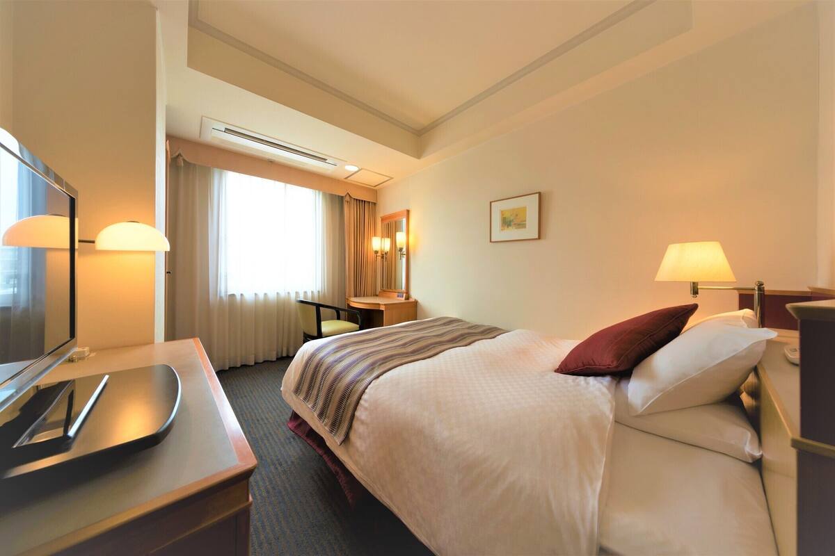 A hotel room with a bed, desk, chair, TV, and a window with curtains.
