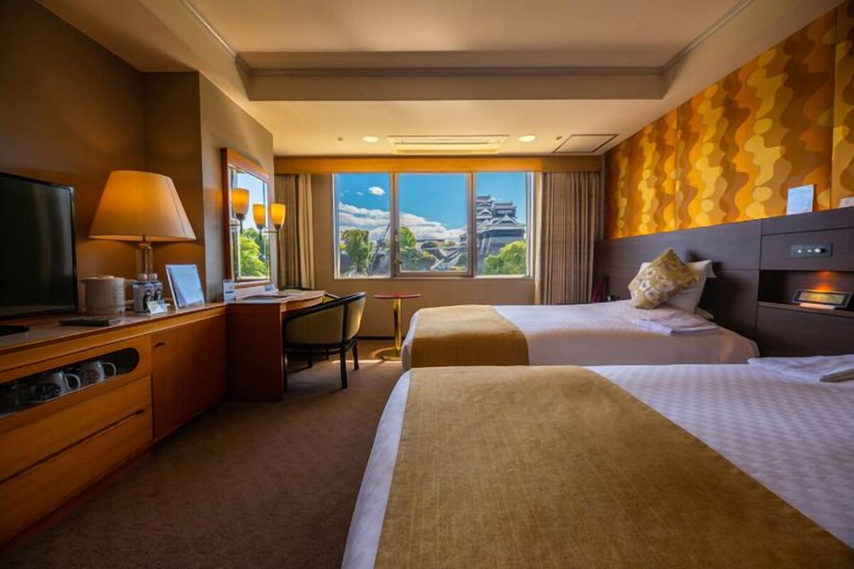 A hotel room with two beds, a desk with a chair, a television, and a window with a view of a building.