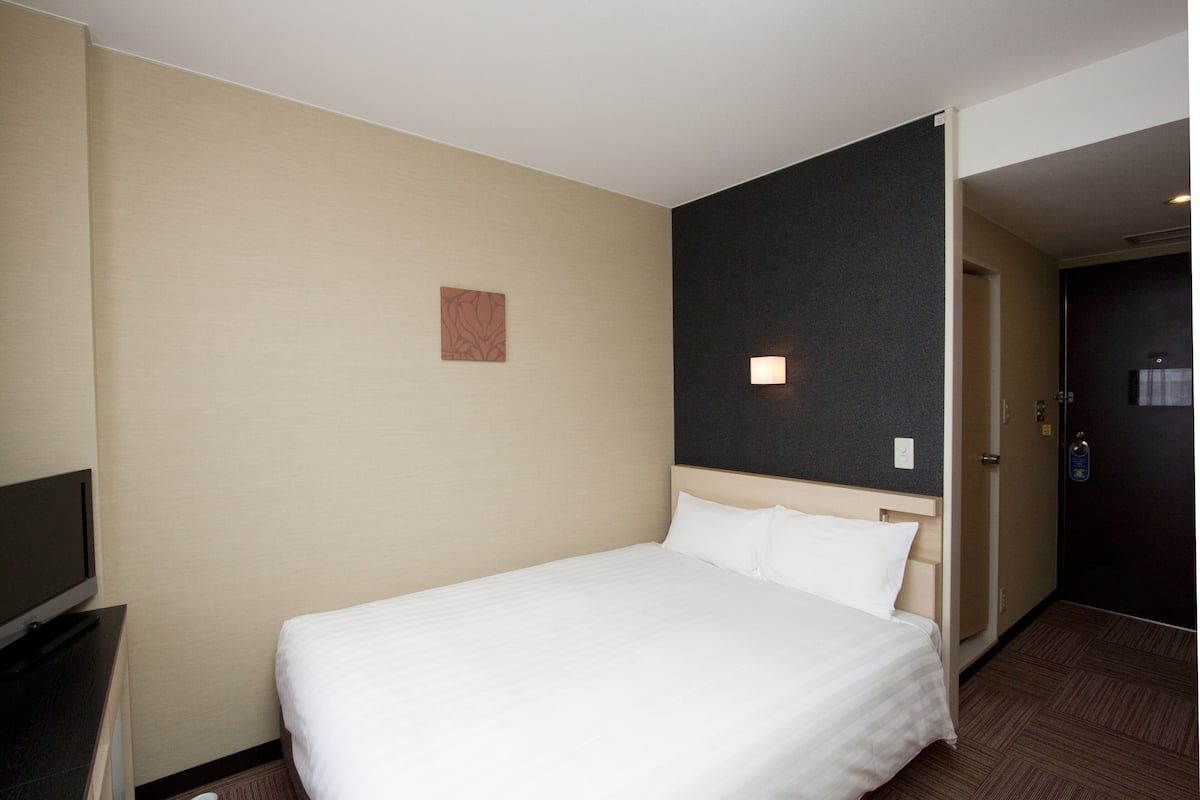 A hotel room with a bed, a television, a door, and a wall with a small artwork.