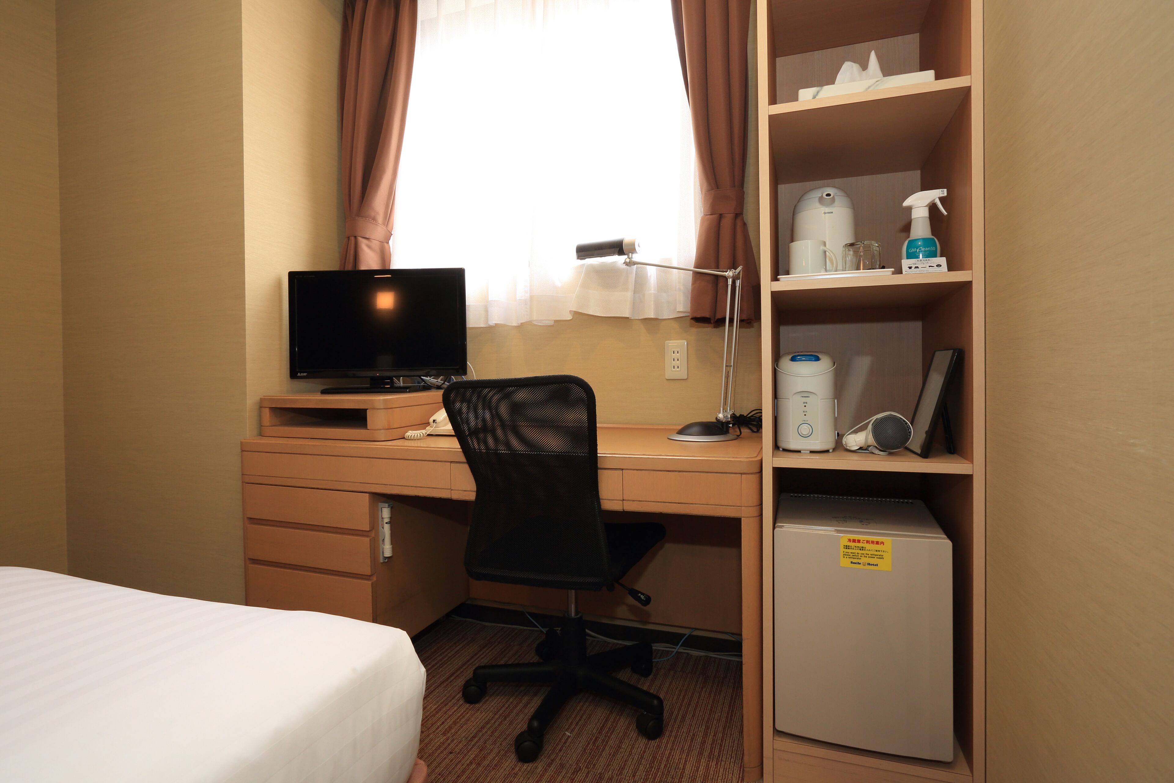 Ein Hotelzimmer mit Bett, Schreibtisch mit Computer, Stuhl, Ablagefläche mit Pflegeprodukten und einem kleinen Kühlschrank.
